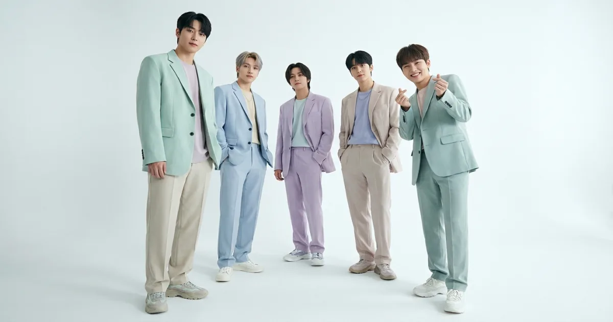 A five-member K-pop boy group standing in formation in coordinated pastel suits, with the youngest member at the far right making a finger-heart gesture — illustrating the formal 'maknae' role in K-pop groups