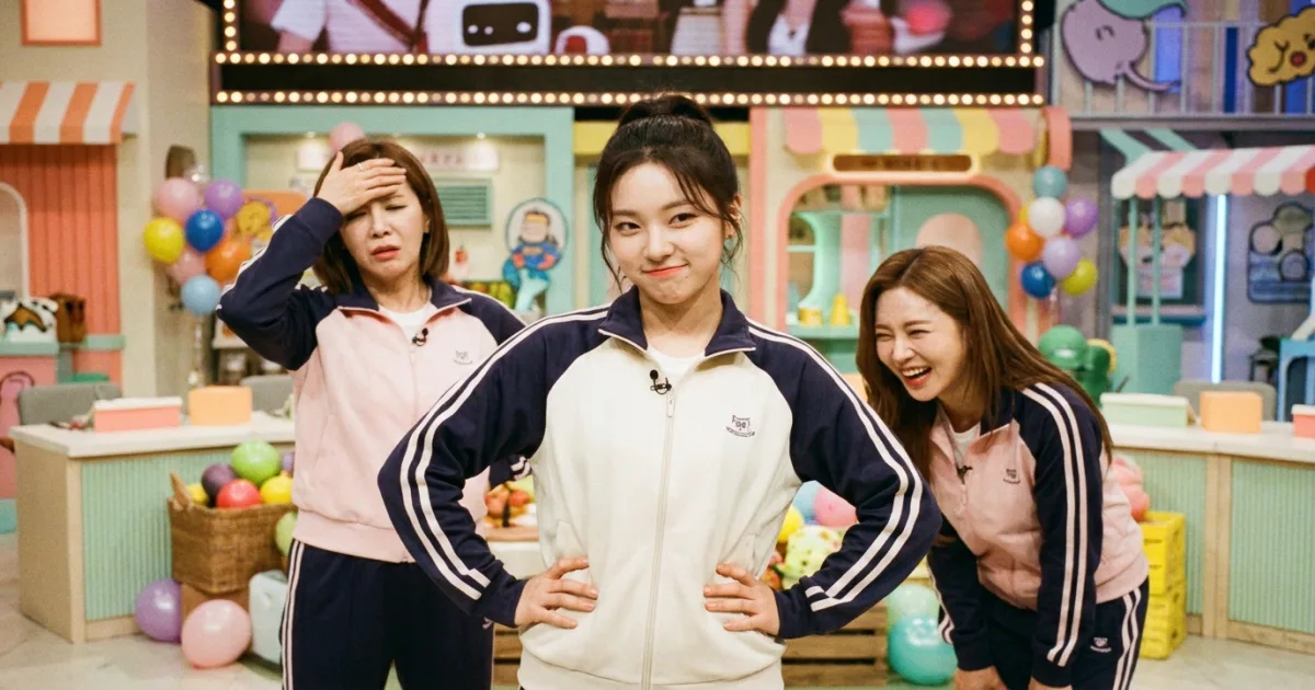 A Korean variety show set with the youngest group member standing confidently in the center with hands on her hips, smirking, while two older members react with feigned exasperation and laughter — the classic 막내온탑 (maknae on top) trope